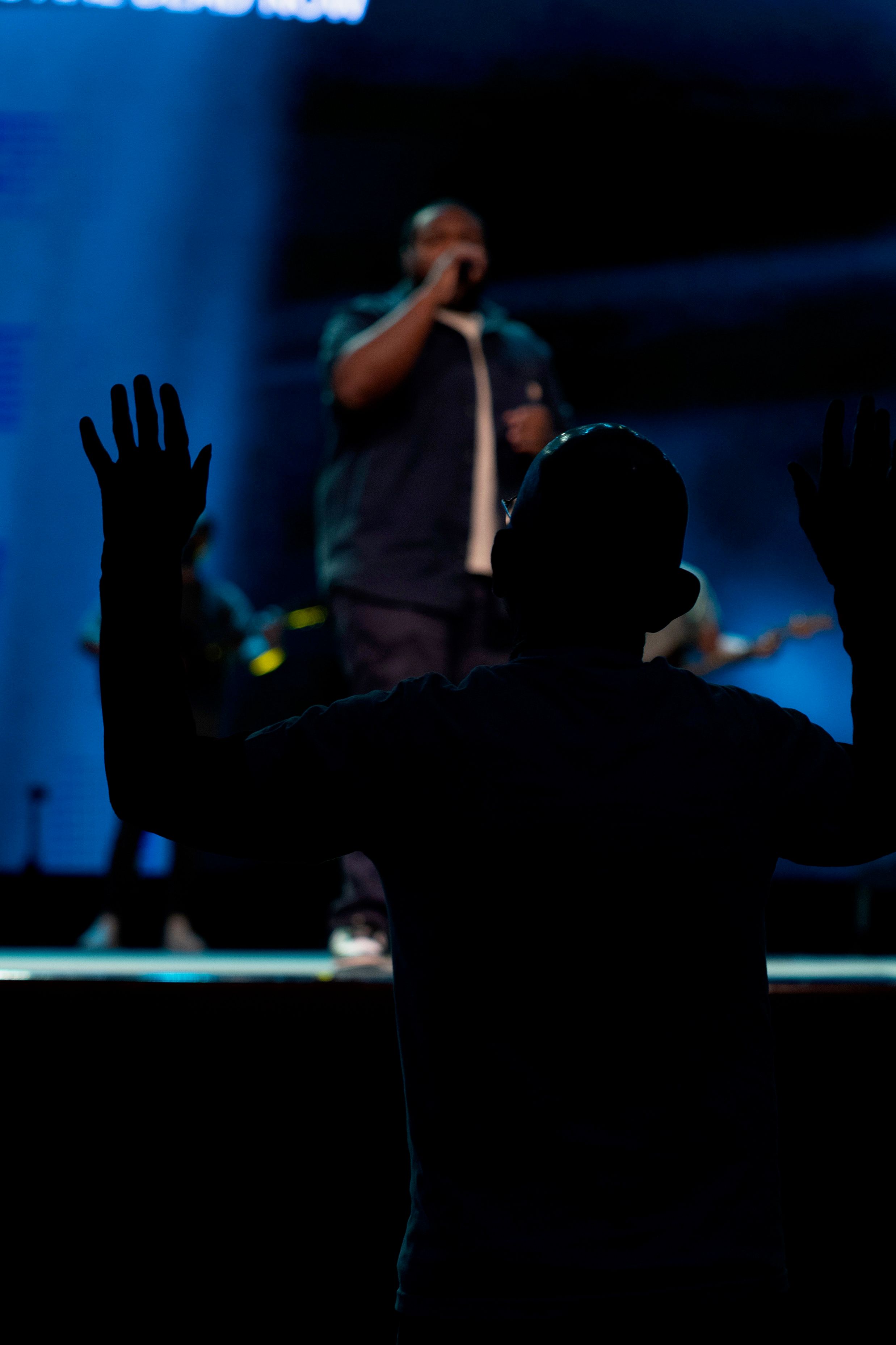 A man seen from behind with both hands raised toward a singer on a blue-lit stage