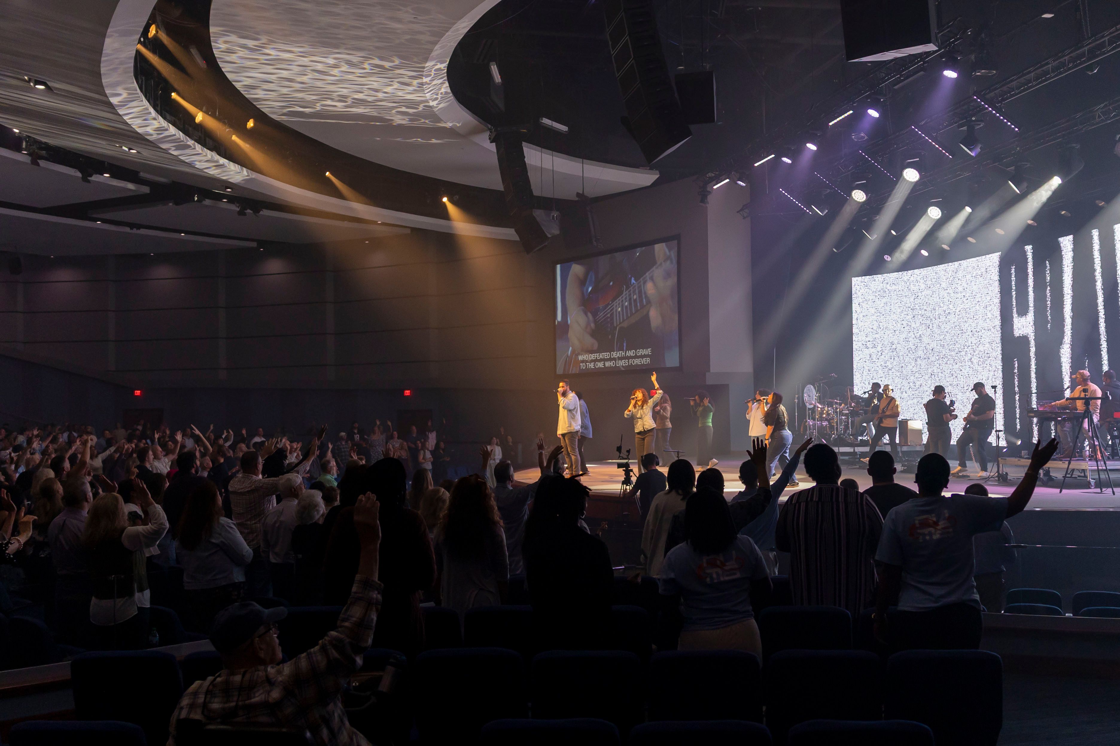 Arena-scale room seen from the back, the crowd dark against a bright stage