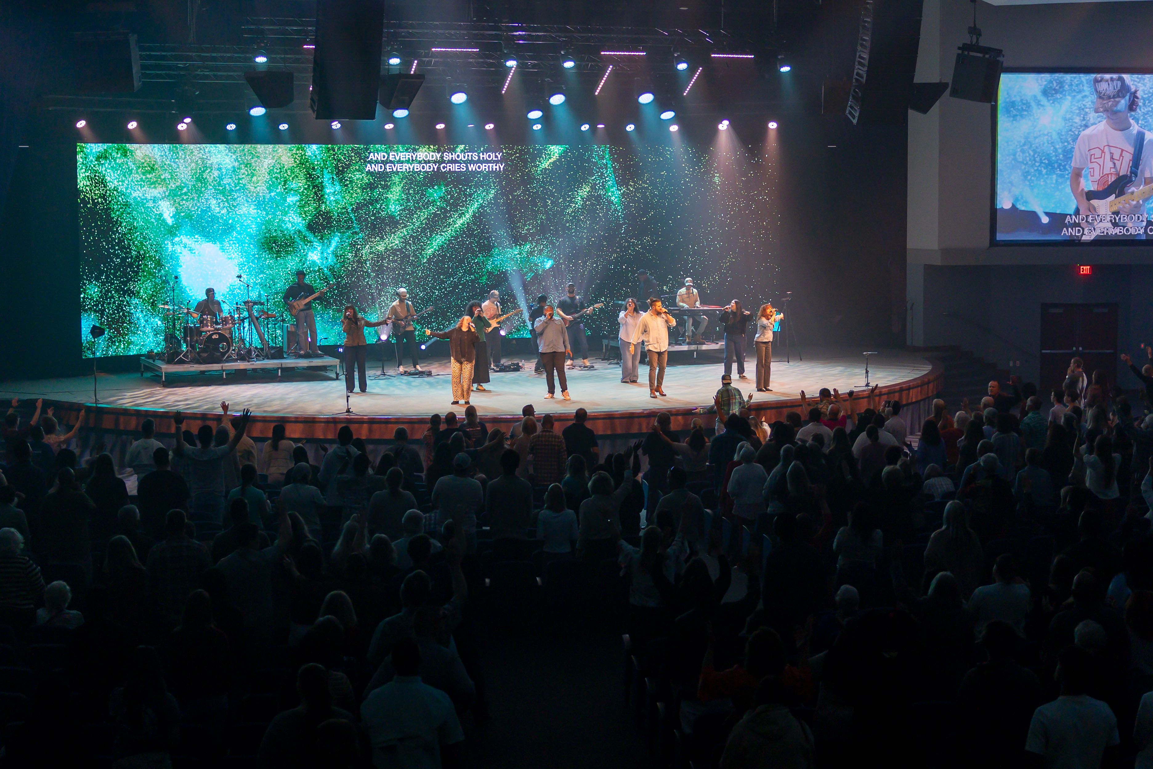 Full worship band spread across a teal stage, lyrics projected above