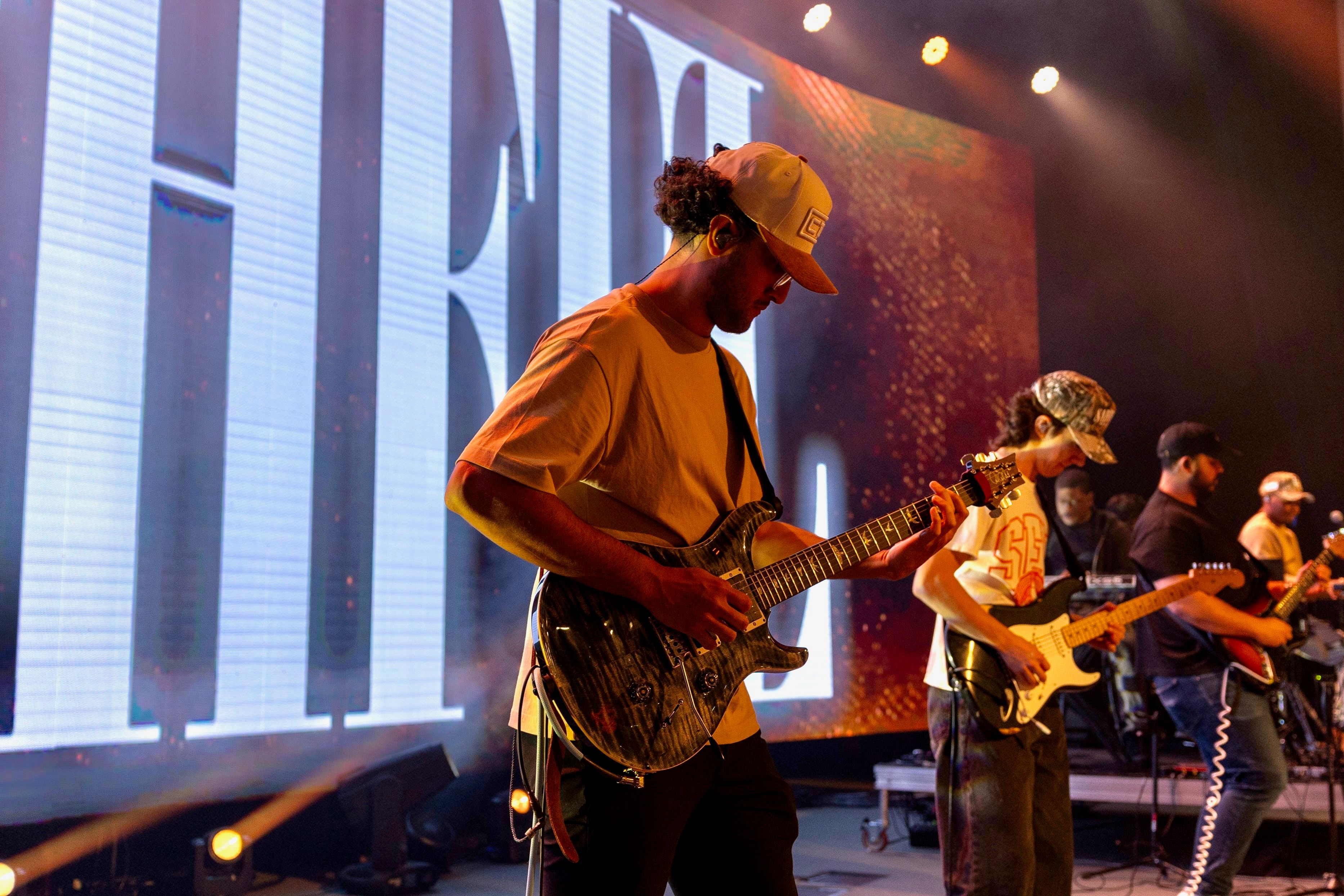 Guitarist in a bucket hat playing beside a bandmate, giant white lettering behind them