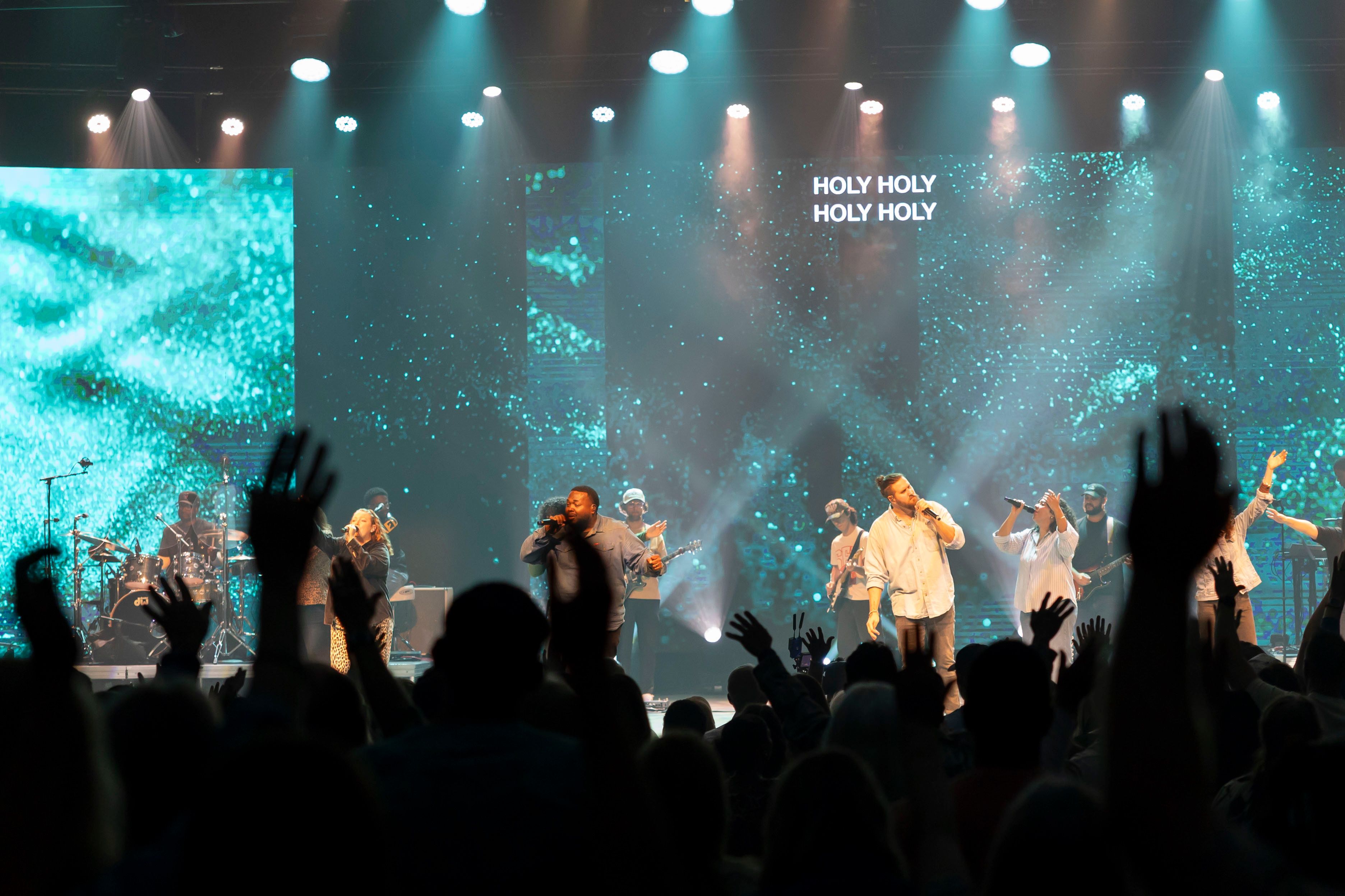 Silhouetted hands raised toward a band on a teal-lit stage