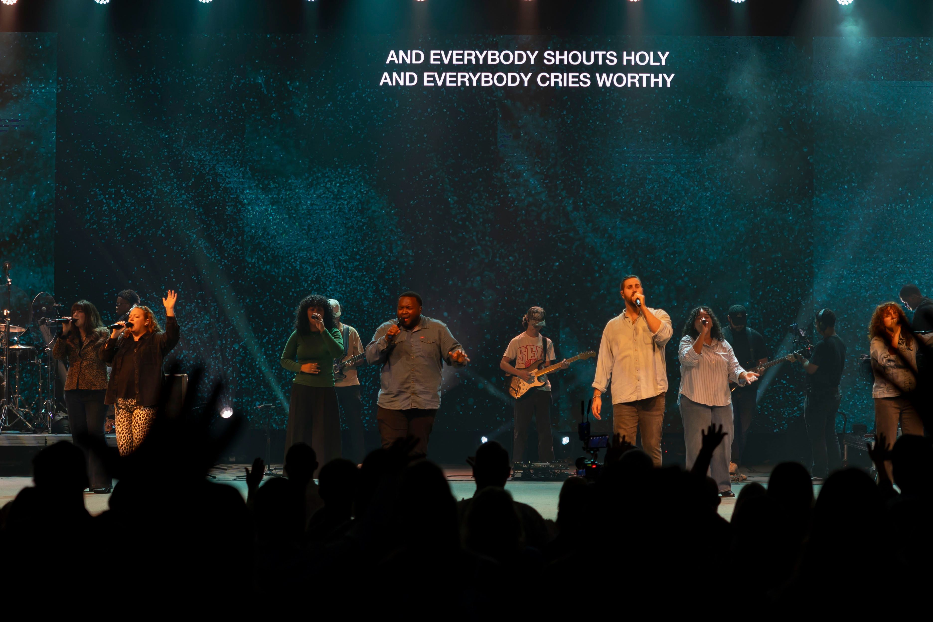Band performing beneath a lyric screen reading "And everybody shouts holy"