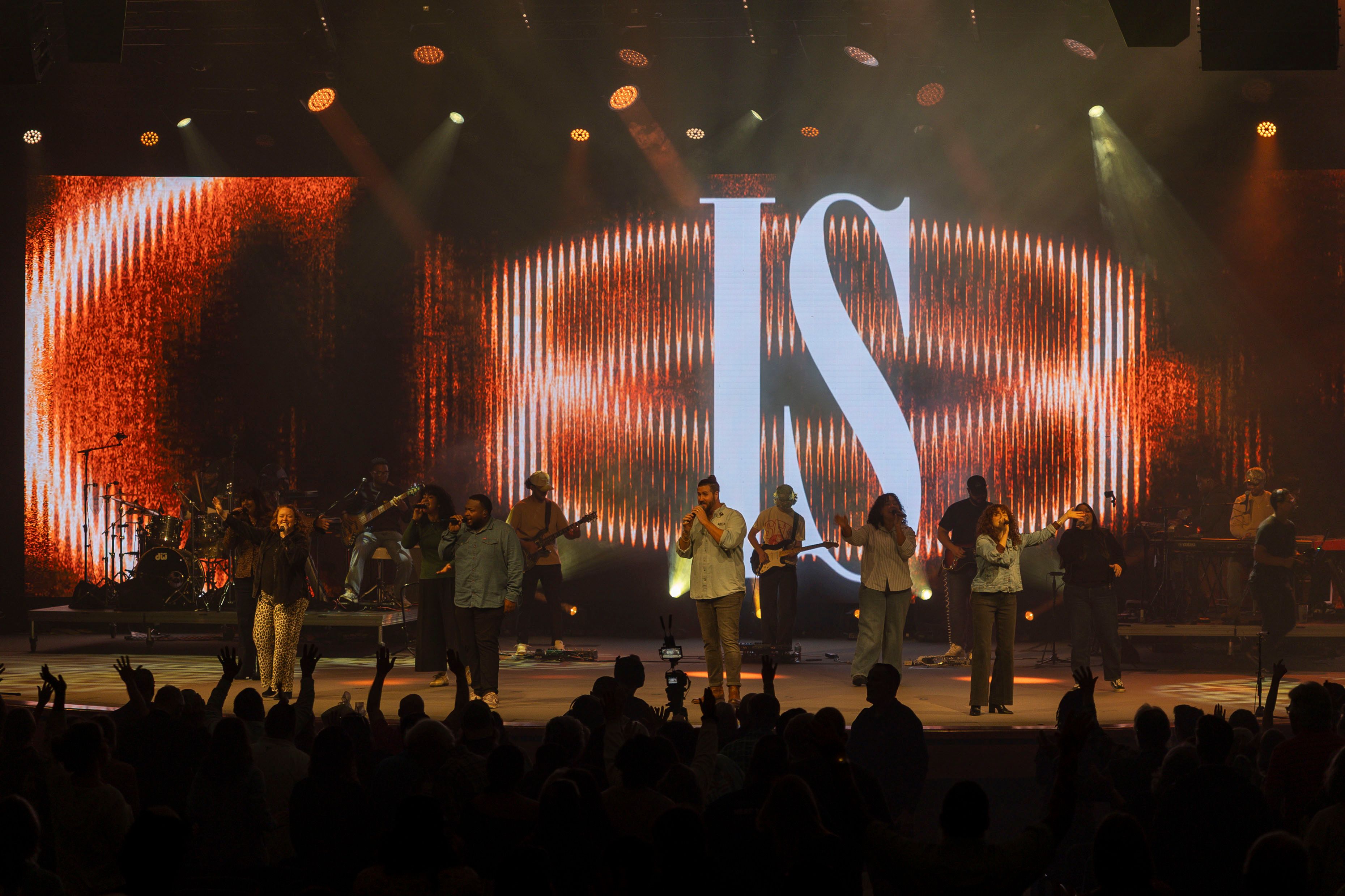 Full band across a warm-lit stage in front of an LED wall spelling "IS"