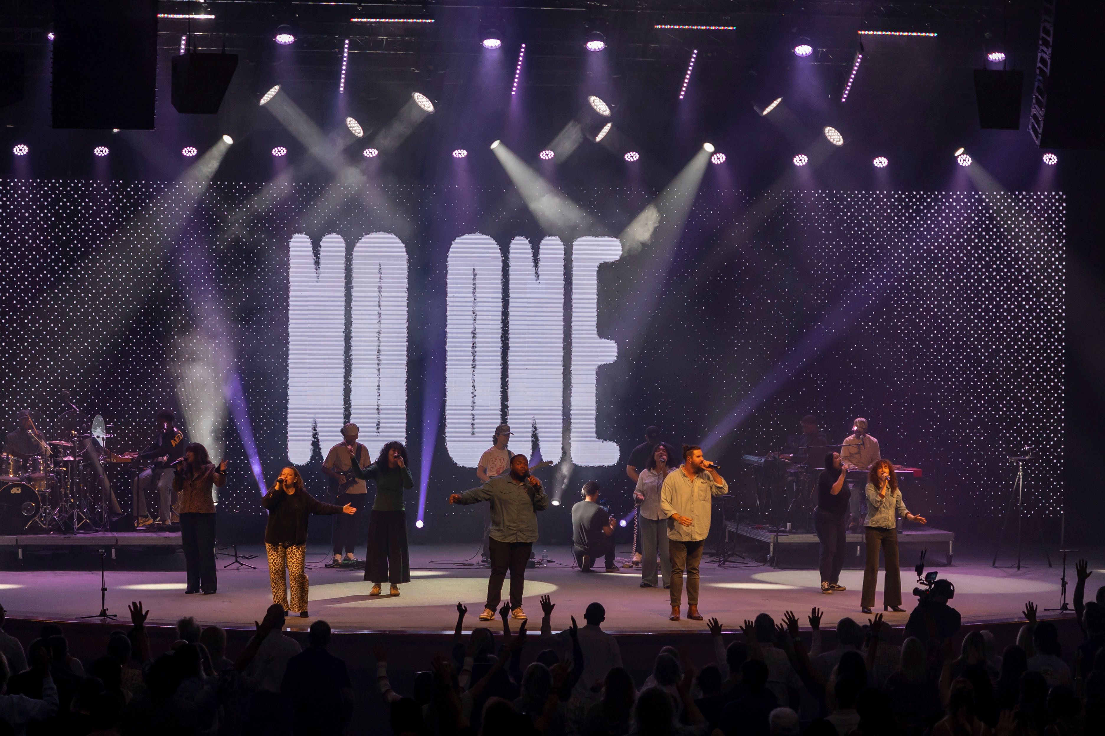 Singers lined across the stage beneath a towering "No One" lyric screen and violet beams