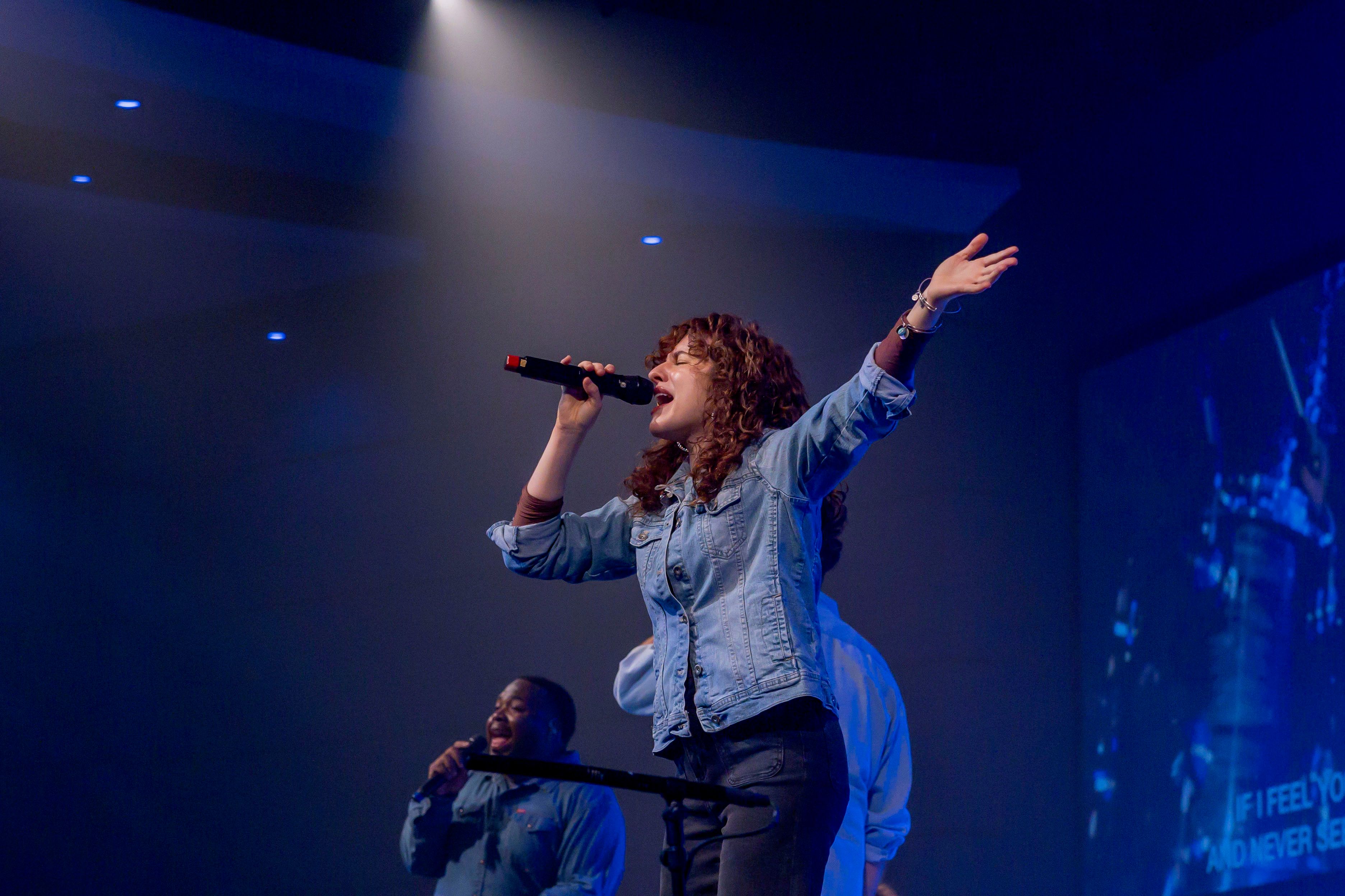 Vocalist with arms open wide singing on a blue stage while a second singer leans in below
