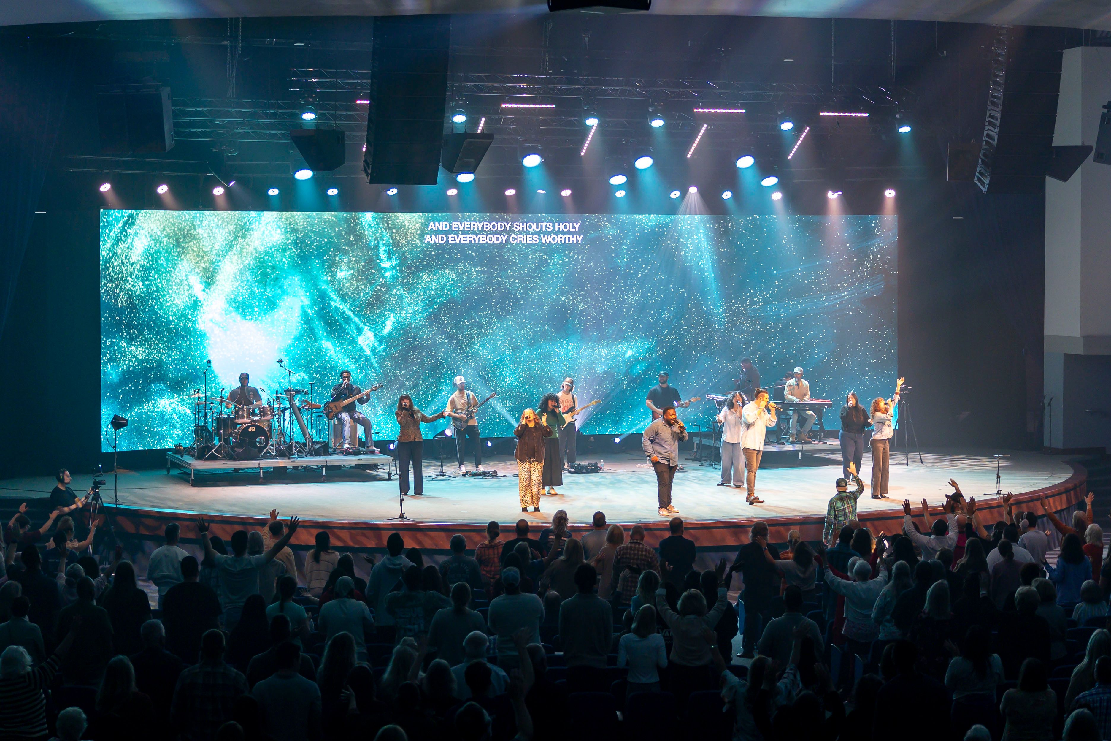 Wide view of a worship band on a round stage washed in teal light, with a packed crowd in the foreground